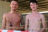 Three young shirtless men stand behind red tape in an outdoor shed. One is holding a beer.