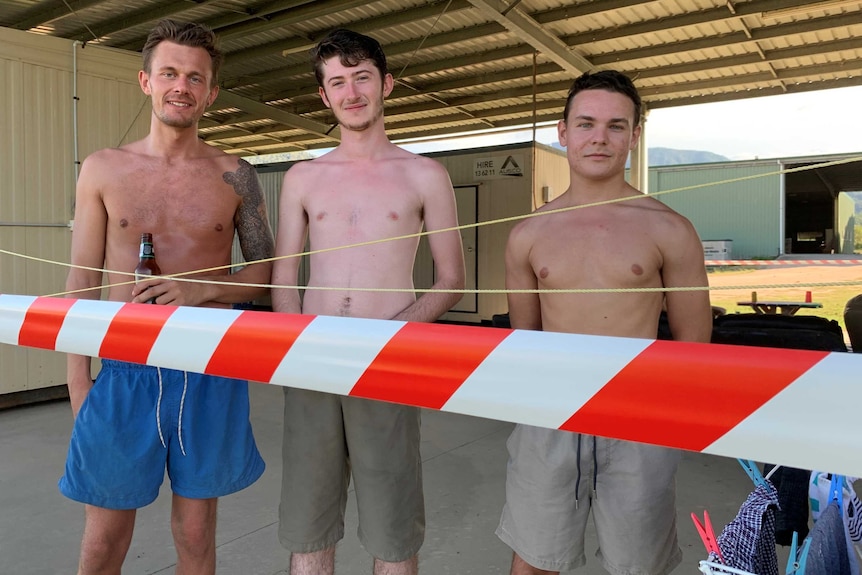 Three young shirtless men stand behind red tape in an outdoor shed. One is holding a beer.