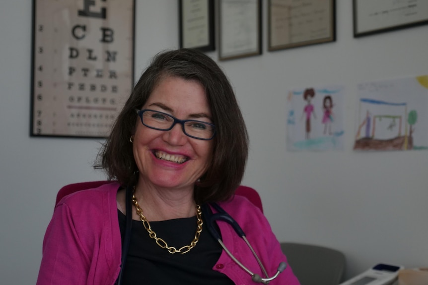 A doctor with children drawings and medical certificates on the wall behind her smiles at the camera