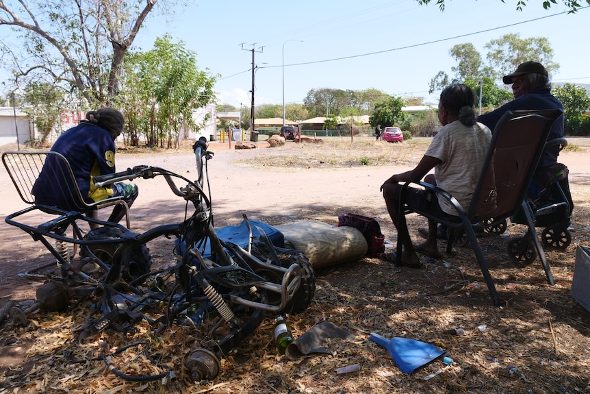 Three older Indigenous people sit on chairs next to a broken bike