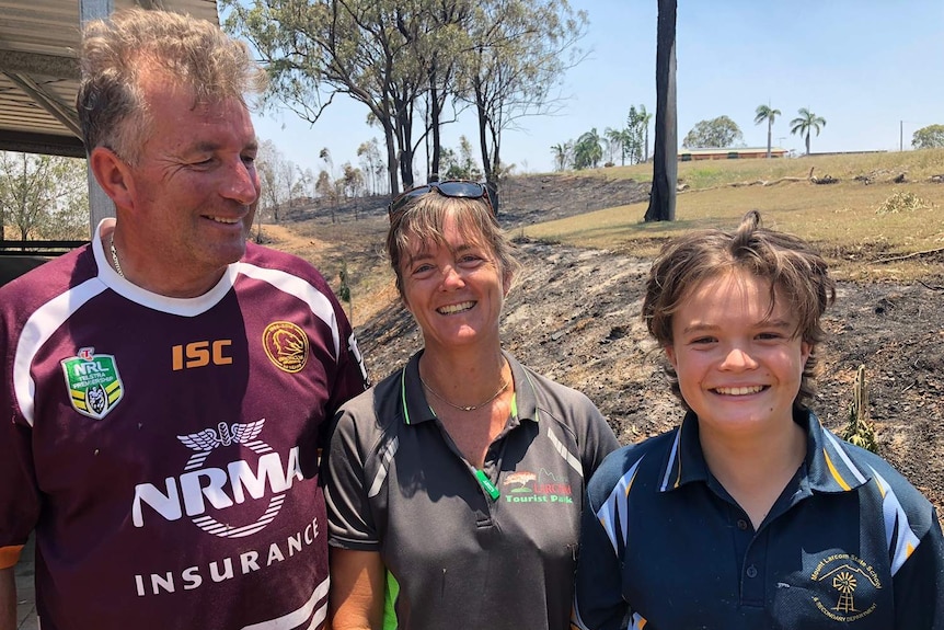 Kev and Del Smaldon with their 13-year-old son Tom smile with burnt bushland behind them.