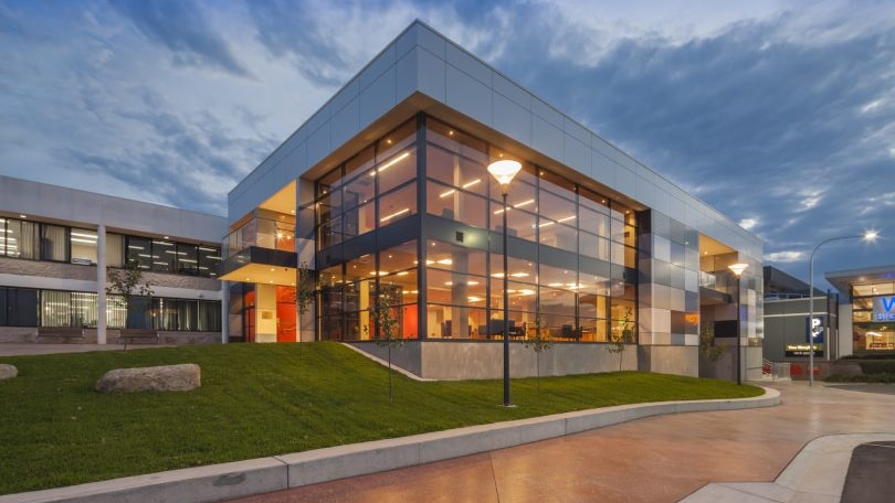 A modern building with lots of glass lit up from inside at dusk with a lawn at its front