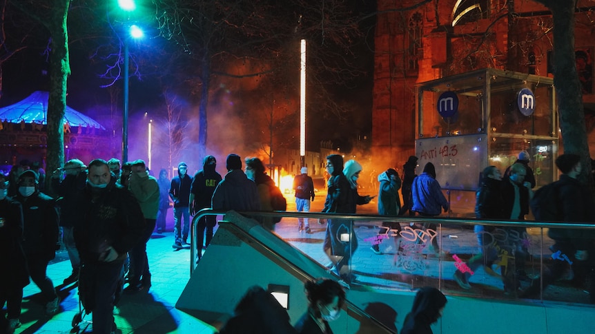 During a demonstration multiple people are pictured walking around as fire burns in the background.