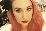 A selfie head shot of Olivia Mead with bright red lipstick, pink hair and a black flower pin in it.