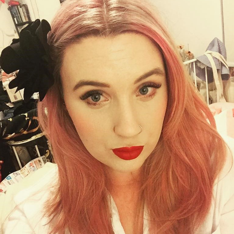 A selfie head shot of Olivia Mead with bright red lipstick, pink hair and a black flower pin in it.