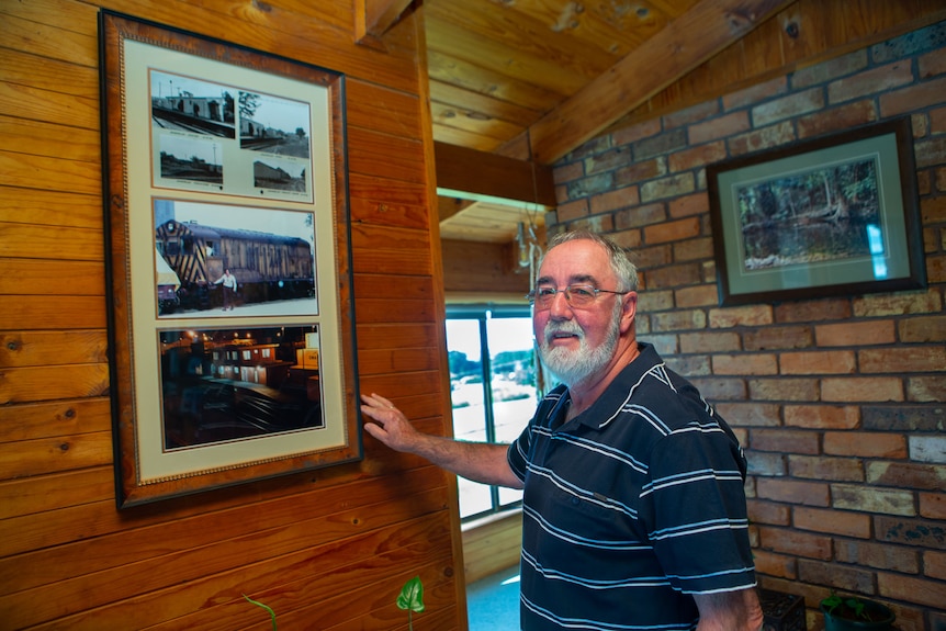 A man stands by a framed selection of train photos in his lounge room.