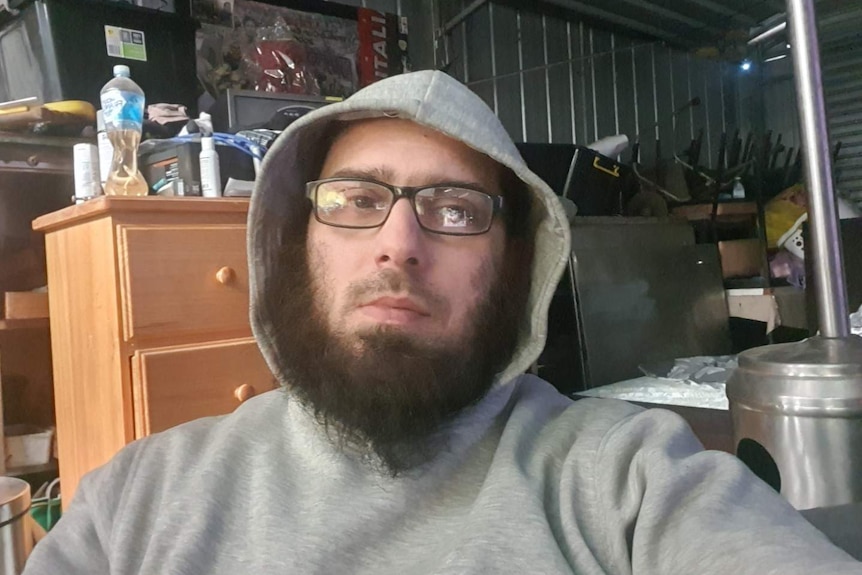 Joe Galliano with a beard, wearing a grey hoodie and black rimmed glasses sitting in a shed filled with odds and ends.