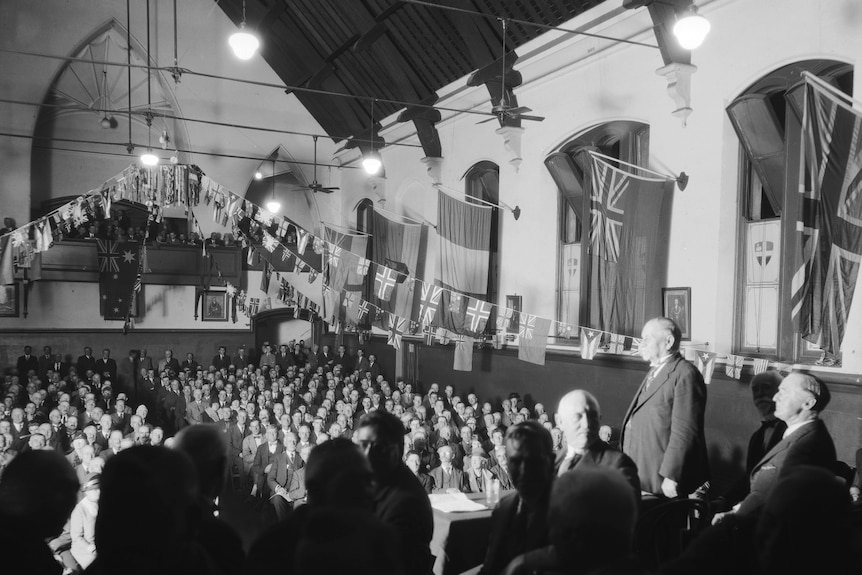 Black and white photo showing men in suits sitting on a stage talking to a crowd of men seated in the audience.
