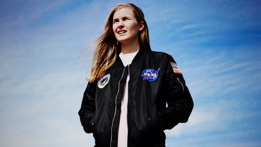 A 2017 press shot of Alex The Astronaut wearing a NASA flight jacket and staring into the sun against a blue sky