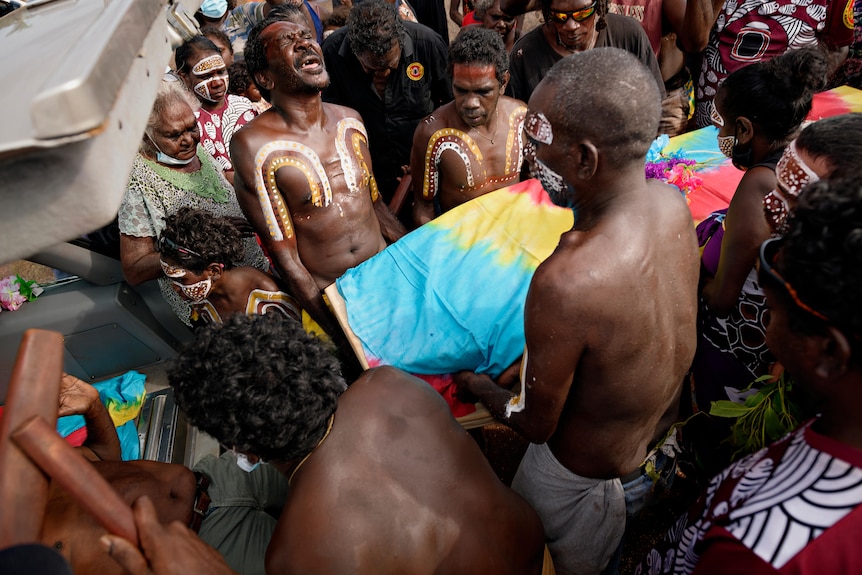 men covered in Indigenous artwork cry and cover a coffin