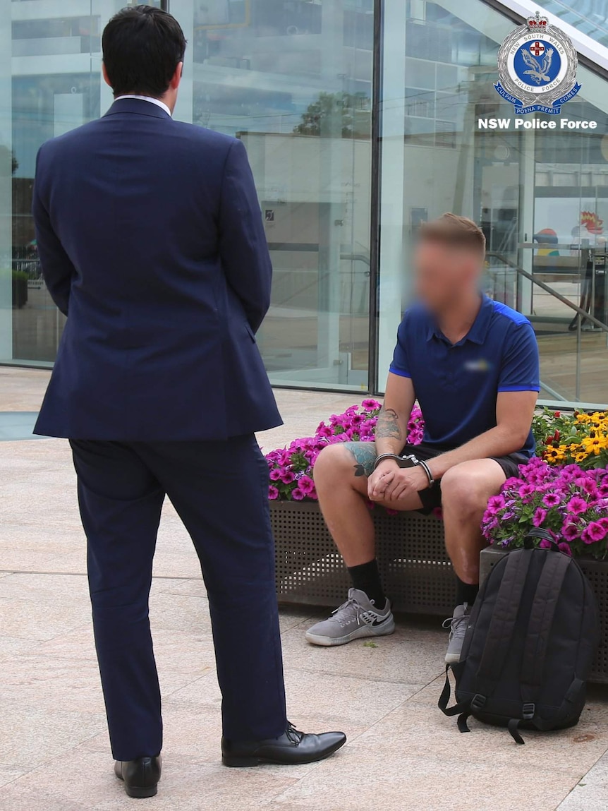 A man in handcuffs sitting in a flowerbed while a man in a suit looks on