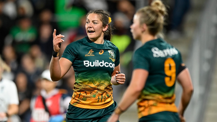 A woman in green and gold football gear shouts instructions to her teammates.