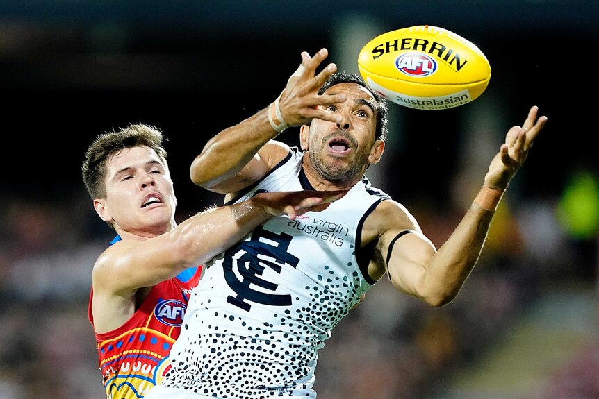 Eddie Betts holds his hands out and looks at a yellow AFL ball while being challenged by a Suns player
