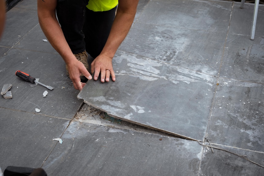 A man lifts up a large grey tile on a rooftop balcony.