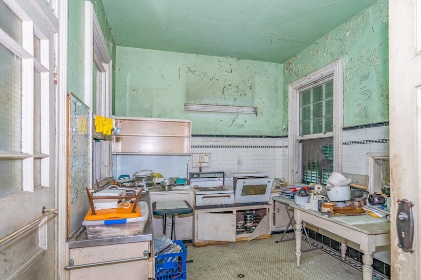 A run down kitchen with old appliances, dishes and paperwork strewn across the benches.
