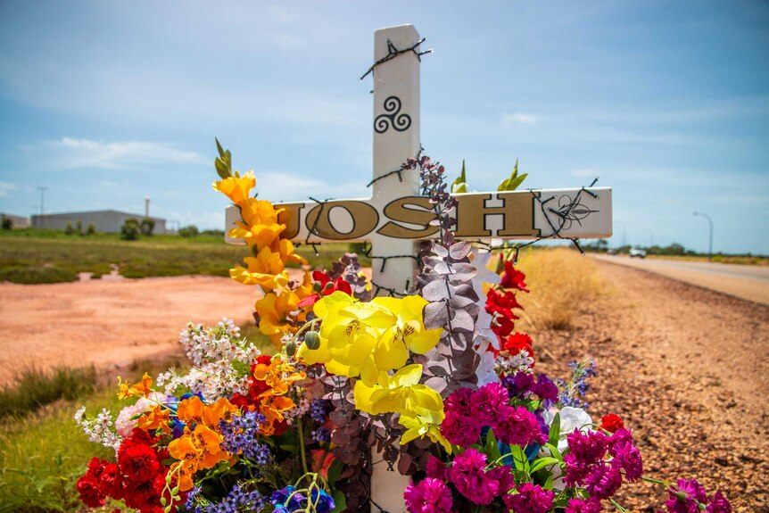 Image of a commemorative cross sitting at the side of a country highway, surrounded by flowers.