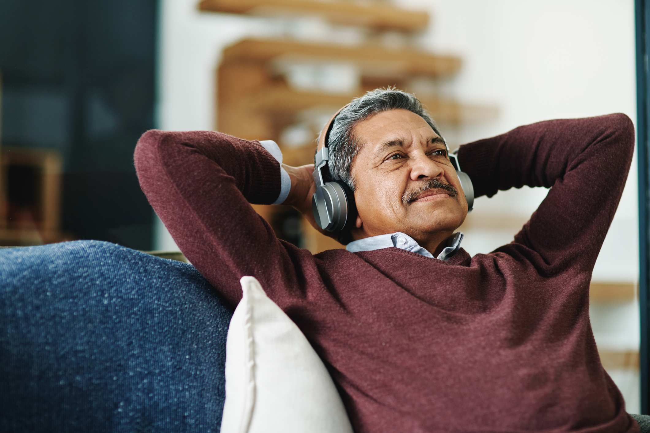 An older Brown man with grey hair and a red jumper leans back in a chair wearing silver bluetooth headphones.