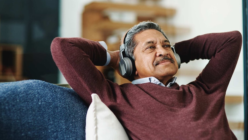 An older Brown man with grey hair and a red jumper leans back in a chair wearing silver bluetooth headphones.