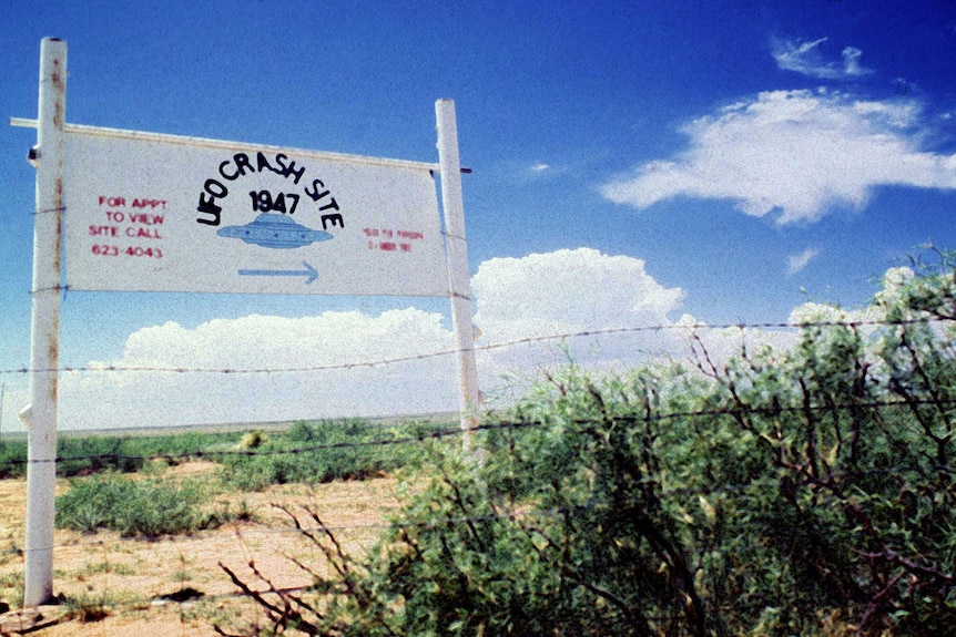 A sign saying UFO CRASH SITE is seen behind a wire fence in the desert