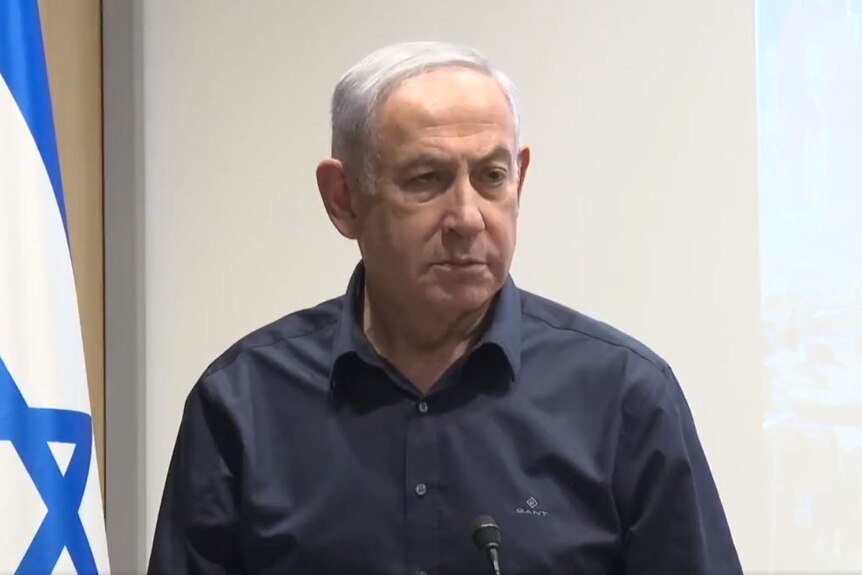 a man with grey hair and blue shirt stands in front of israel flag