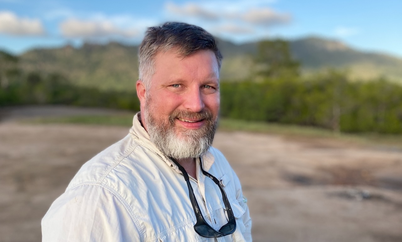 a white man standing in a national park with green bush in the background