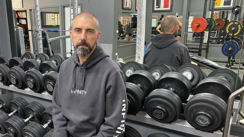 A bald headed, bearded man in grey workout gear sits against a weight rack in a gym.