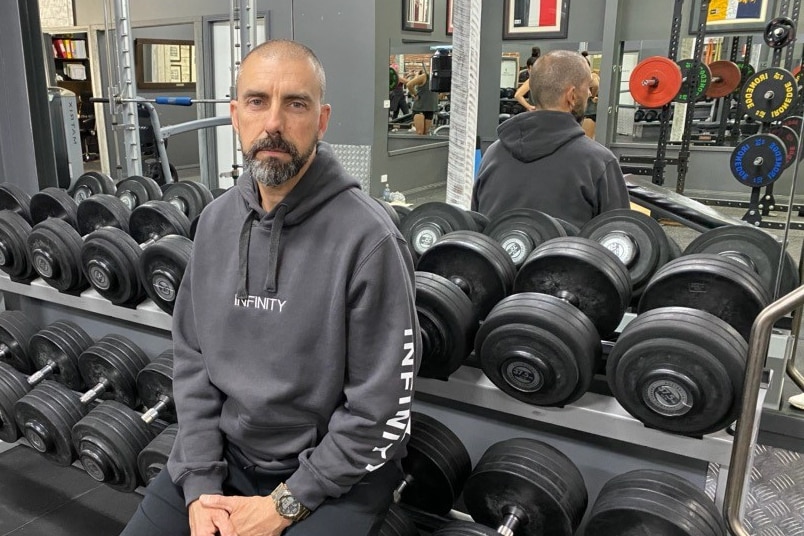 A bald headed, bearded man in grey workout gear sits against a weight rack in a gym.