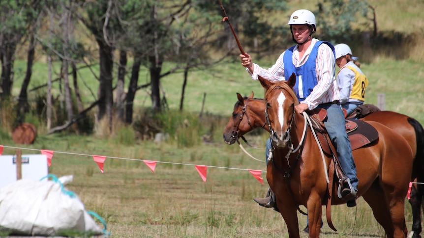 A man in a blue bib and white helmet raises a stock whip while mounted on a horse. 