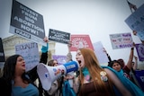 A young woman with long red hair holds a megaphone, while surrounded by other people brandishing placards