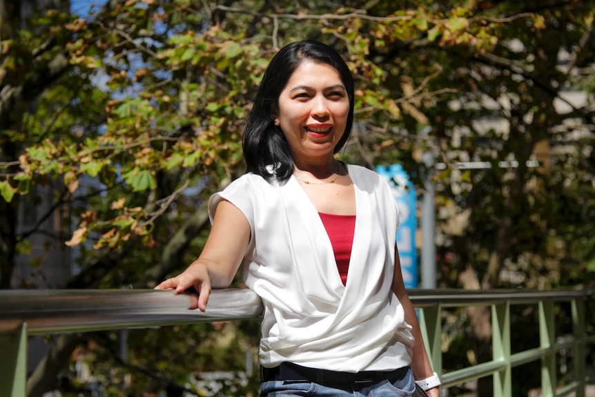 Esperanza- with white shirt and black hair - stands against a railing with trees behind her.