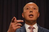 The photo is taken from beneath Mr Dutton, who is standing at the lectern and pointing with his right hand. There are two flags.