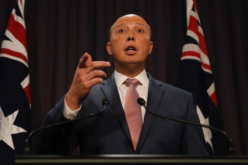 The photo is taken from beneath Mr Dutton, who is standing at the lectern and pointing with his right hand. There are two flags.