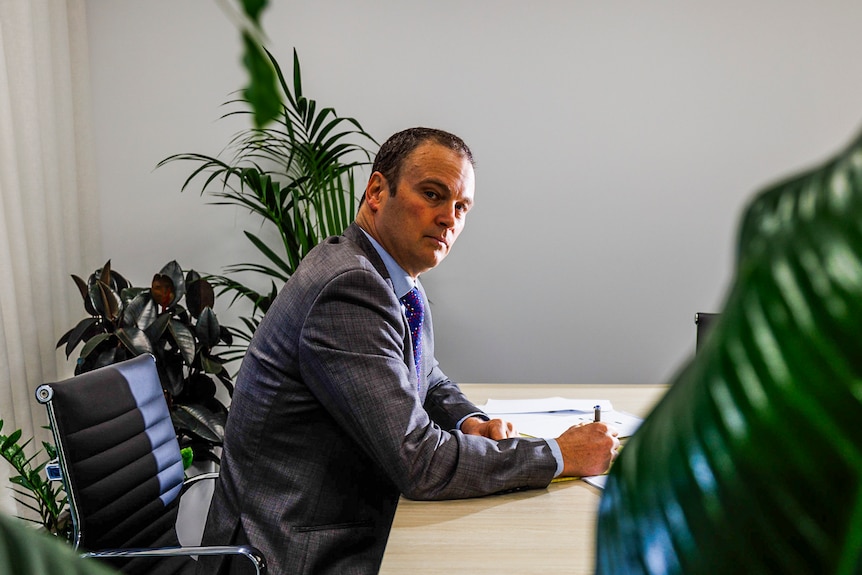 A man in an office sits at a table and turns to face the camera with a serious expression.