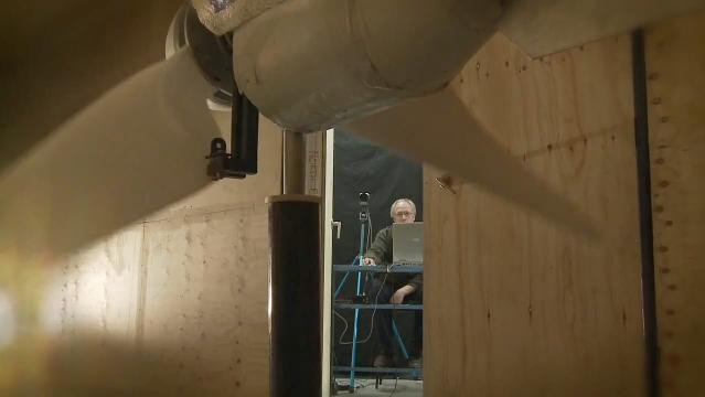 A wind turbine in a laboratory setting, a man sits at a laptop computer in the background