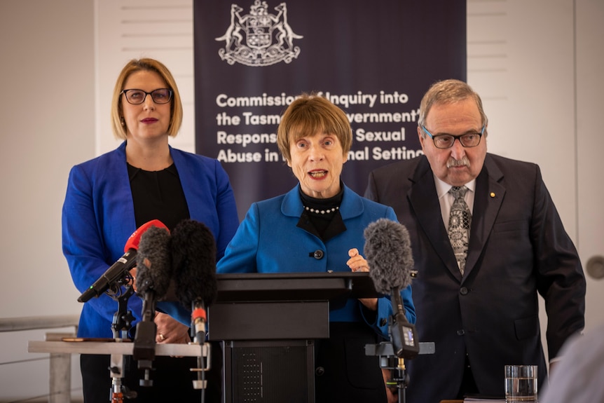 Two women and a man stand at microphoned podium addressing the media 