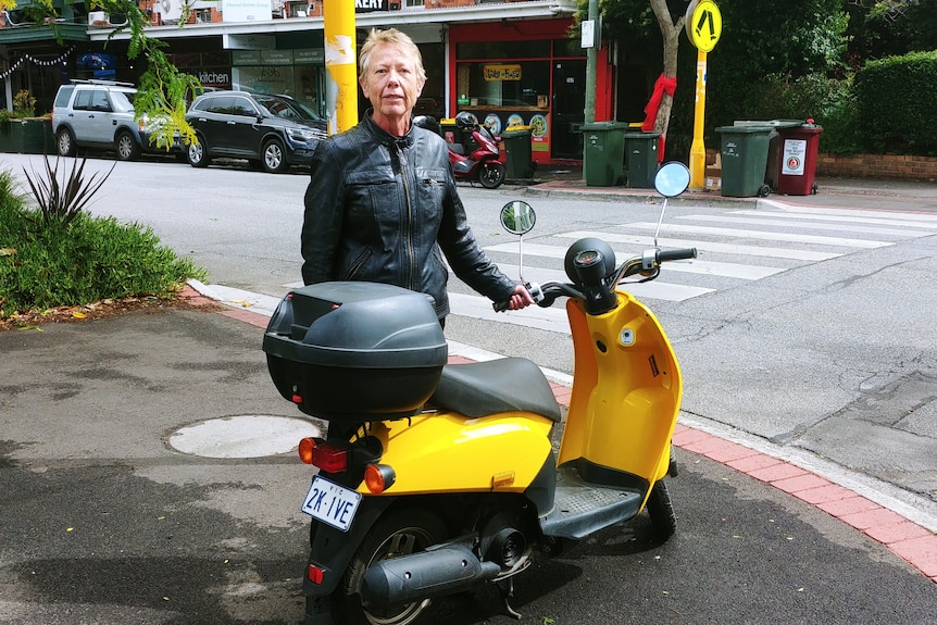 An older woman in leather near a bright yellow scooter on a suburban street