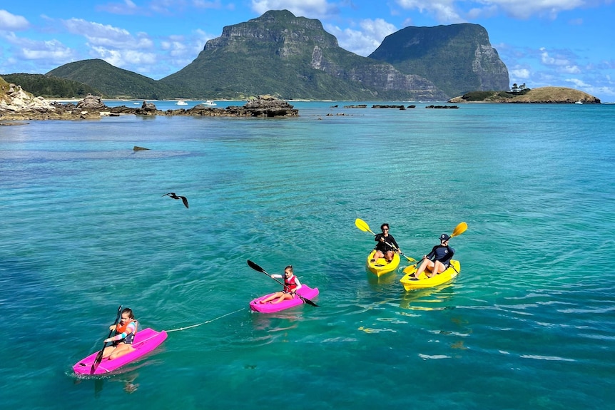 Two adults and two children paddle brightly coloured kayaks on a clear blue island lagoon.