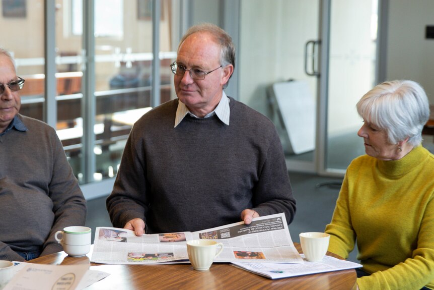 Three people sitting at a table with cups of tea and reading a newspaper