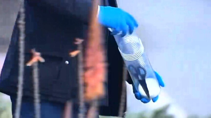A close-up shot of a forensic police officer with blue rubber gloves holding a plastic container with a pair of scissors inside.