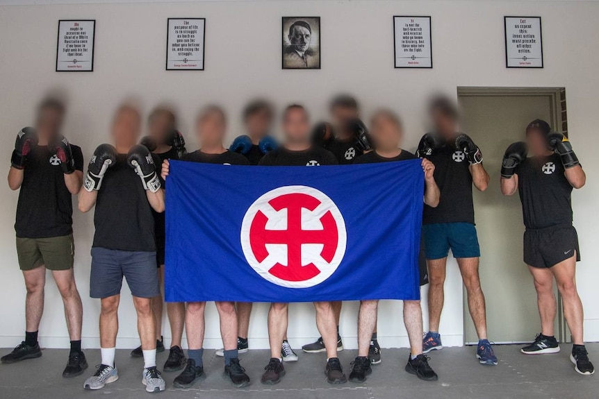 Group of men with blurrred faces stand in front of a portrait of Hitler and holding a flag with a sign and wearing boxing glov
