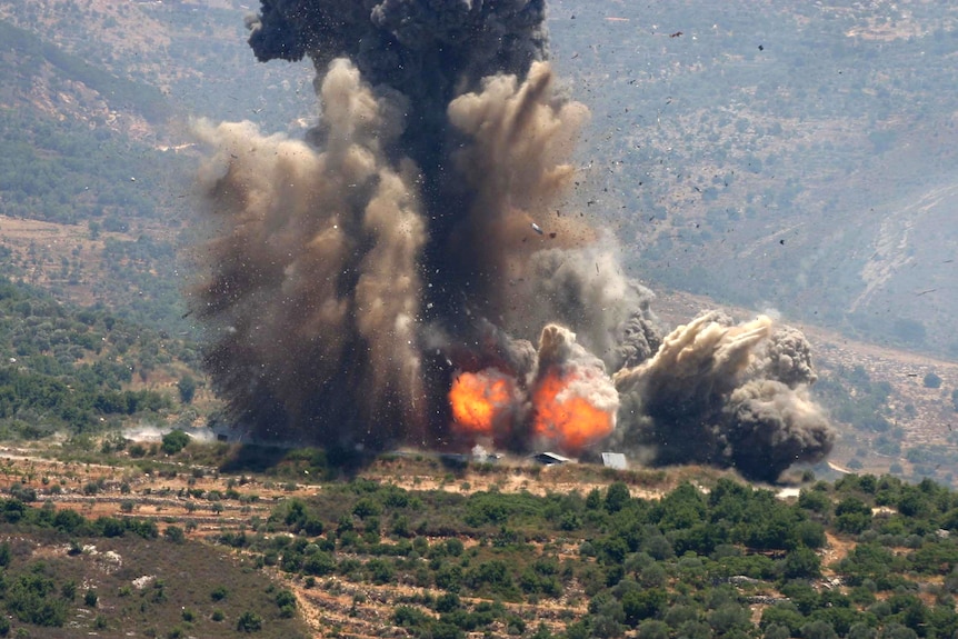 An explosion goes off in the distance, in hilly terrain with several structures visible
