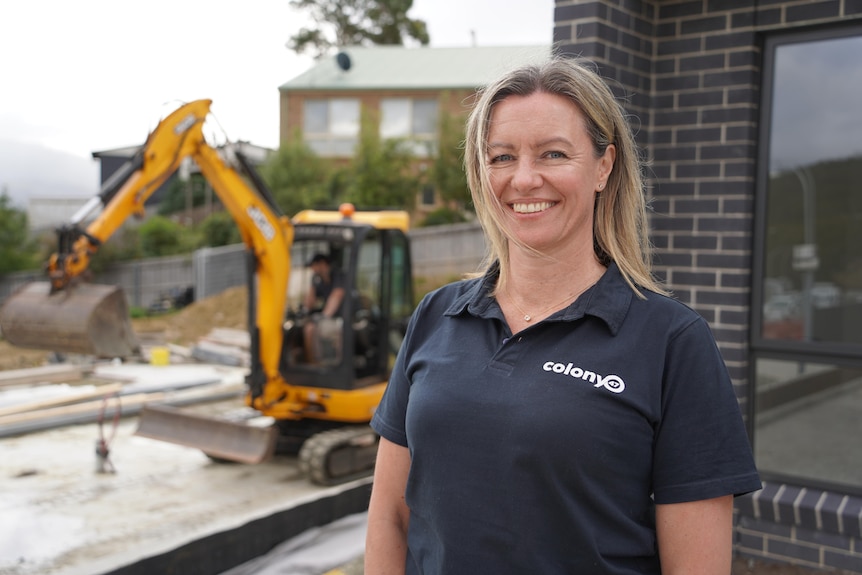 Dianne Underwood stands outside a newly-constructed grey brick home, with a digger visible in the background
