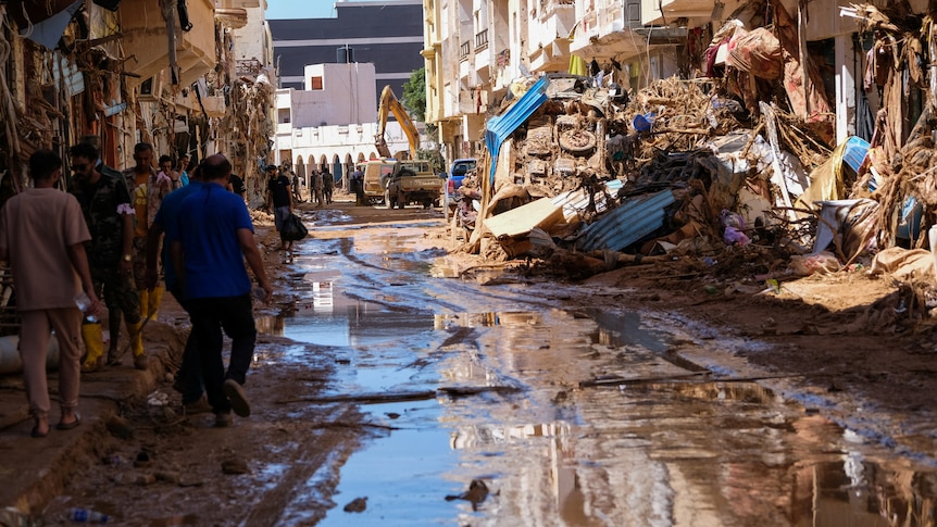 People walk in the mud between destroyed buildings, an excavator is piling up debris