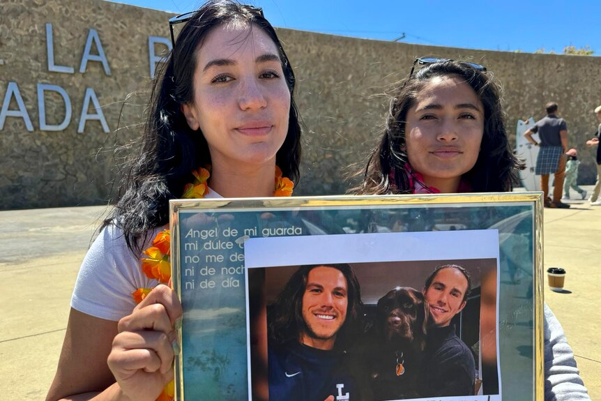 Two women hold a framed photo of Jake and Callum Robinson in front of a large outdoor wall.