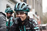 A cyclist wearing black and white leans on her handlebars and smiles to the camera.