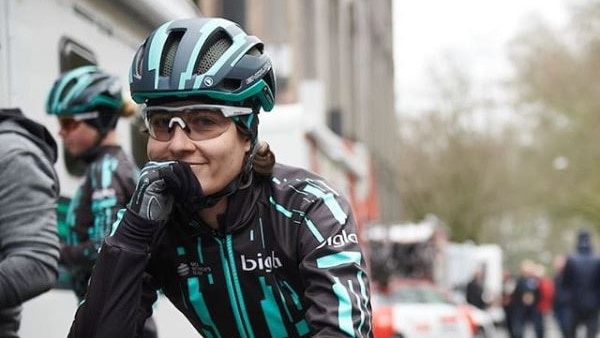 A cyclist wearing black and white leans on her handlebars and smiles to the camera.