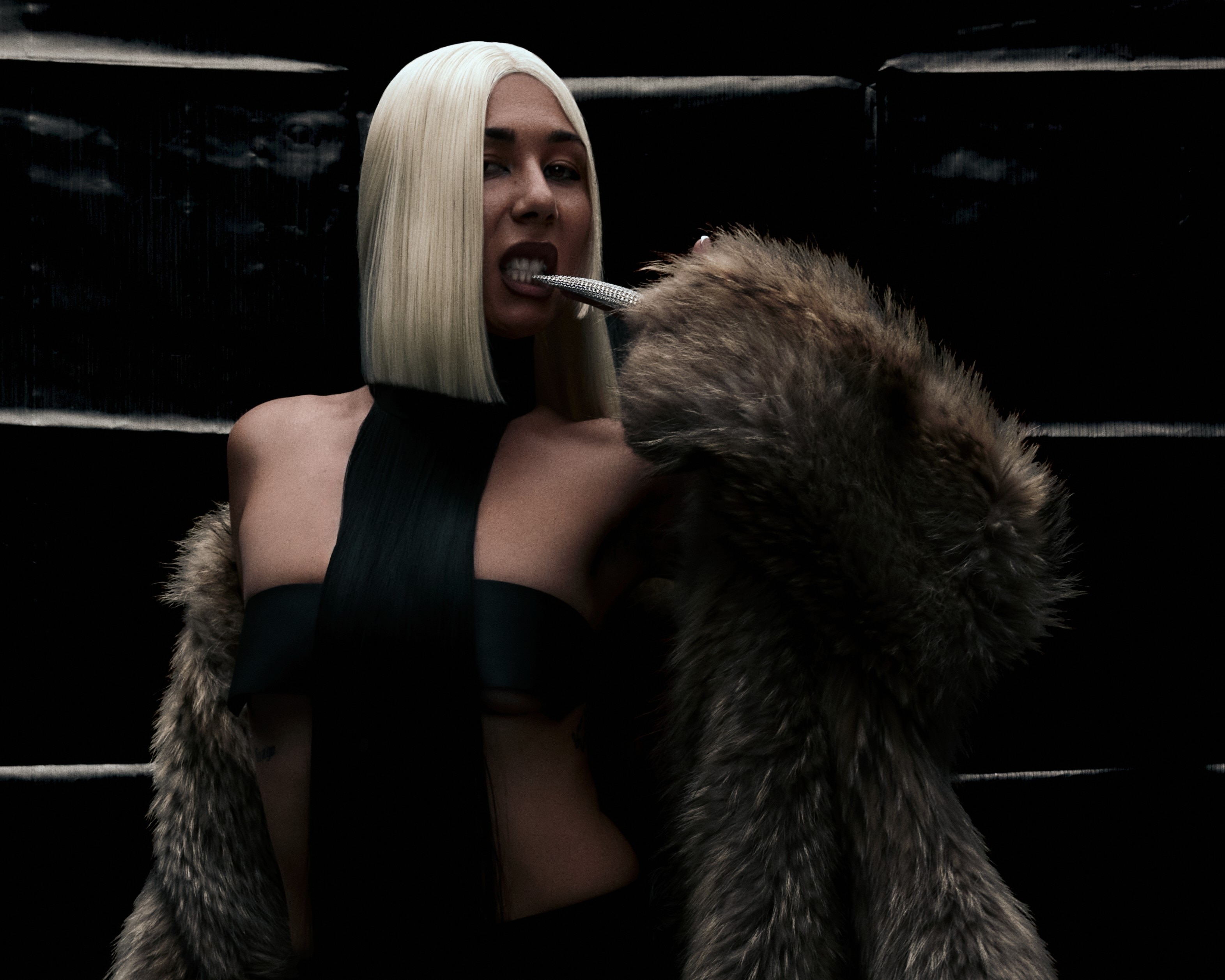 An Indigenous woman with platinum blonde straight hair bites a finger-long silver claw while wearing a fur coat