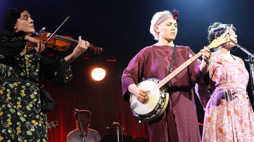 Three women play violin, banjo and sing respectively during a stage production.
