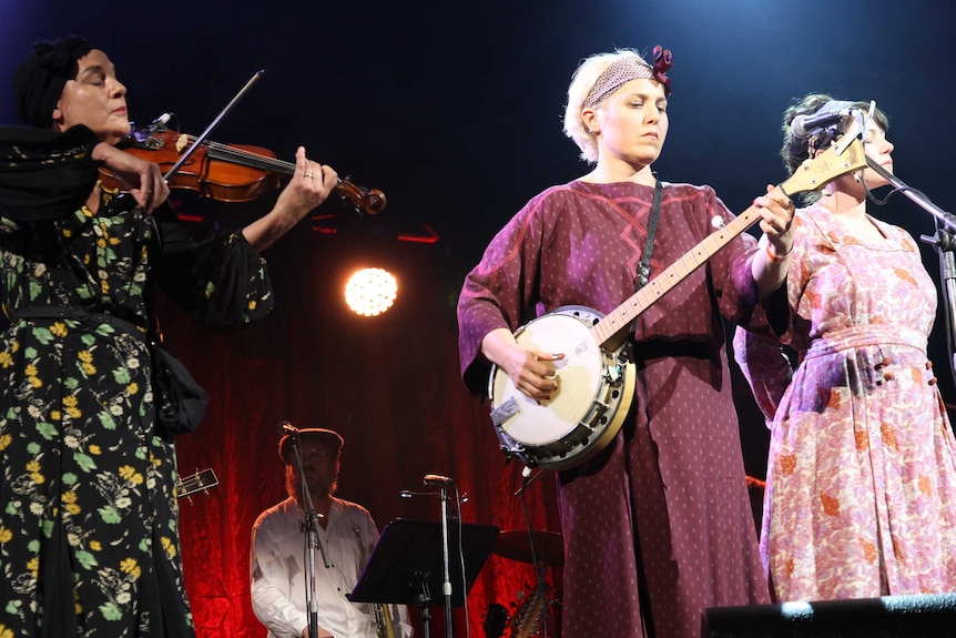 Three women play violin, banjo and sing respectively during a stage production.
