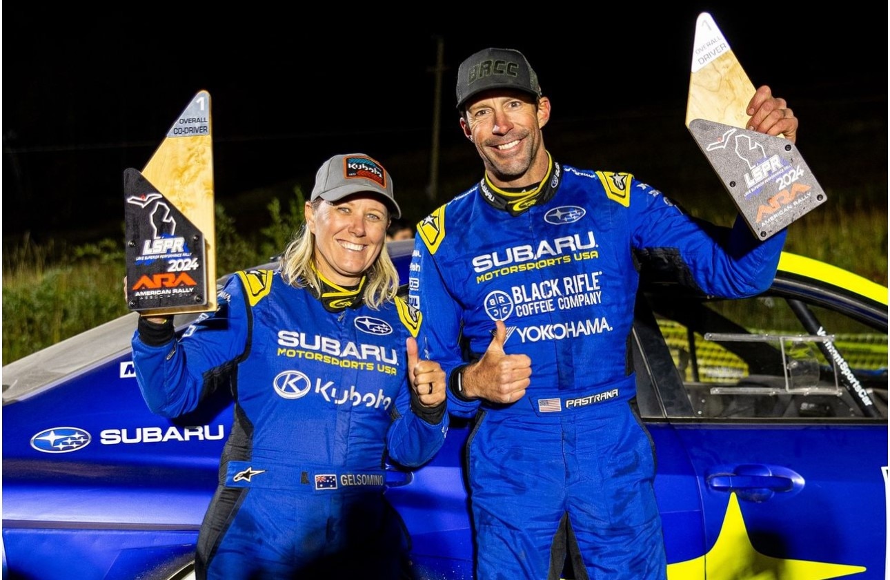 Plavokosa žena i viši, tamnokosi muškarac u trkačkim uniformama koje stoje ispred plavog automobila koji drži trofeje.